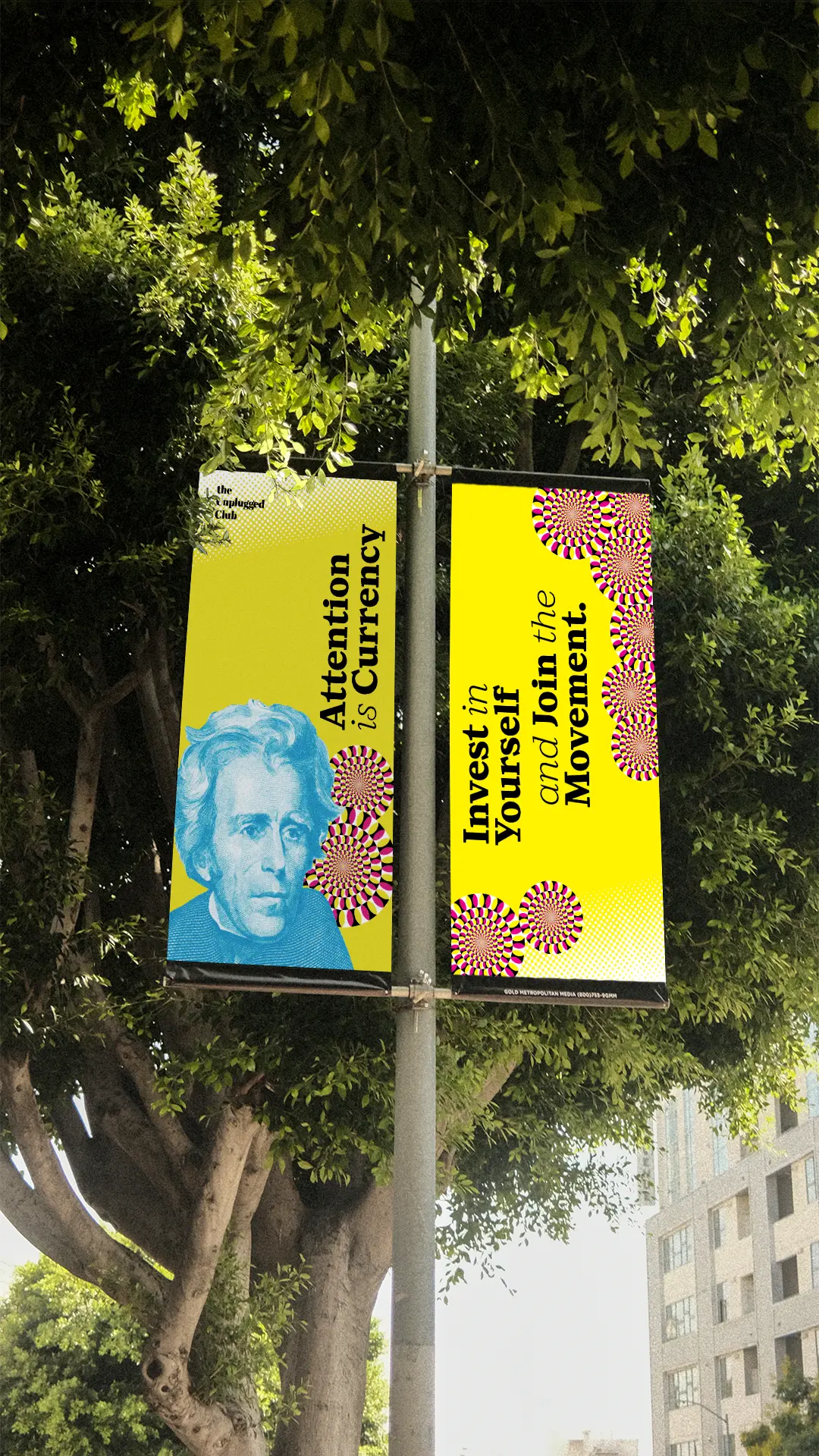 A pair of banners, hanging froma street pole in front of a tree. Both of the banners are yellow. An image of Andrew Jackson in cyan and the copy in black color are printed. The copy says, 'Attention is Currency. Invest in
Yourself and Join the Movement.